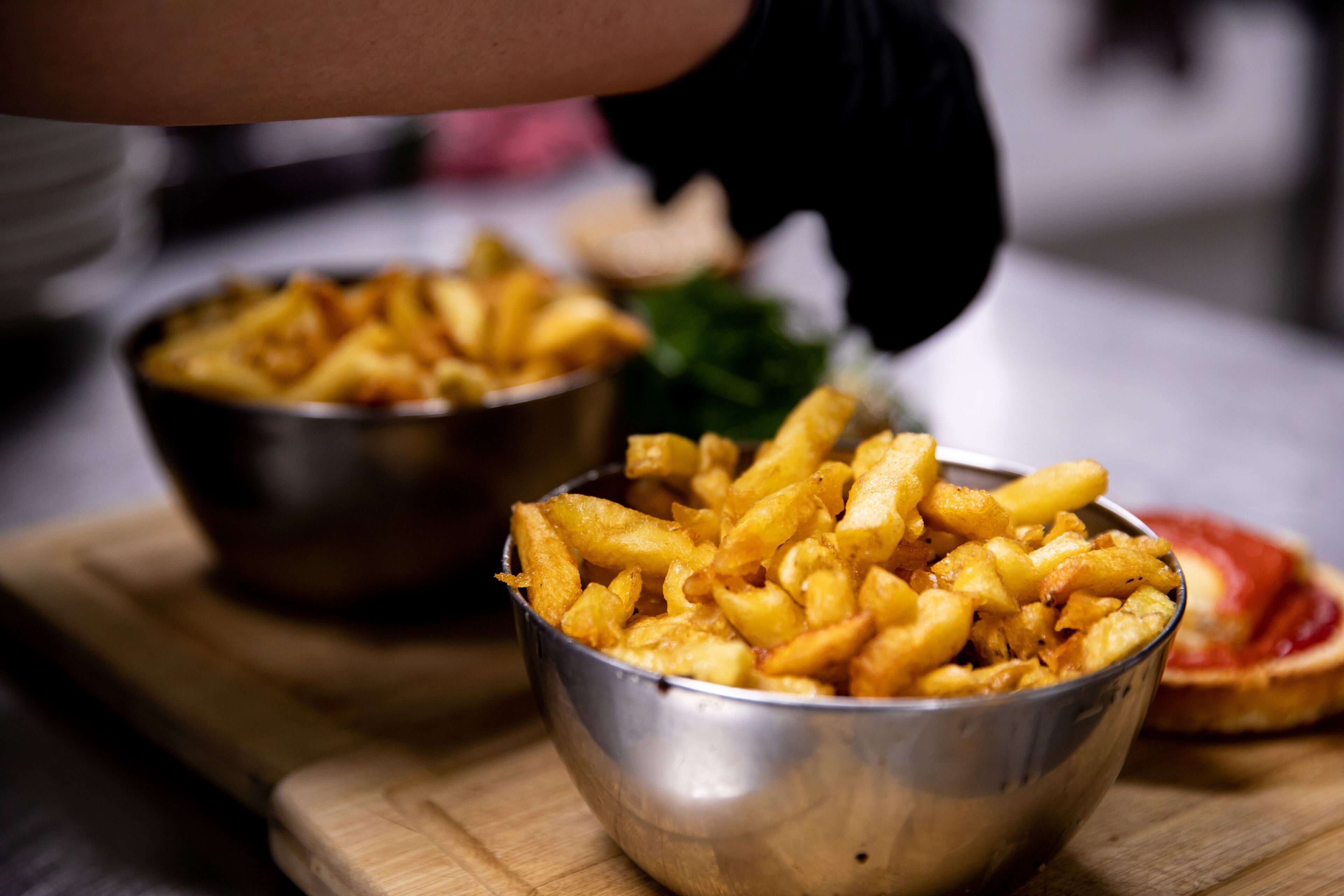A metal bowl filled with golden fries on a wooden surface, with another bowl and a slice of bread in the background.