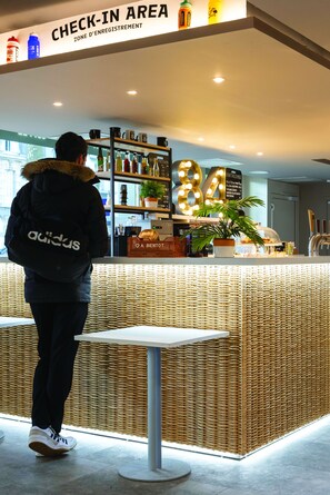 Un área de recepción con un cartel que indica 'CHECK-IN AREA' y 'ZONE D'ENREGISTREMENT'. Hay estantes con varios artículos, una planta en maceta y un cartel con el número '84'.