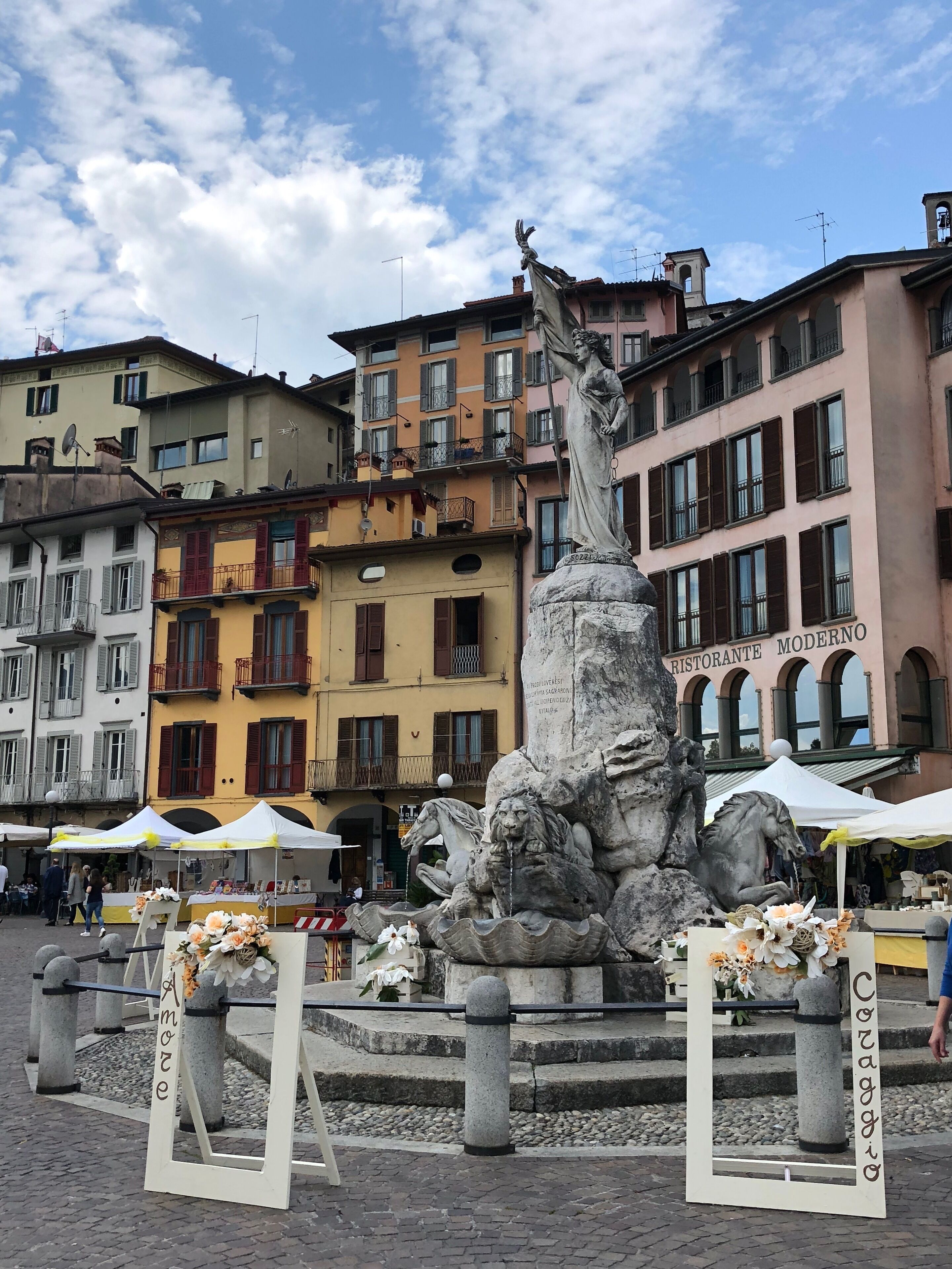 A public square with a fountain, surrounded by historic buildings and outdoor market stalls.