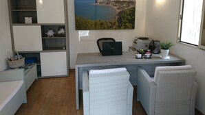 Un espacio de oficina compacto con un escritorio, dos sillas, una estantería, un aire acondicionado de pared y un ventanal con vistas a un paisaje costero.