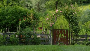 A rustic wooden gate with a metal archway adorned with pink flowers, set in a lush green garden with a wooden fence and trees in the background. - The Music Country House, between Asolo, the Prosecco hills and Monte Grappa (Castelcies)