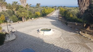 A paved courtyard with a central fountain, surrounded by greenery and a pathway leading to a distant view. - Korisia accommodation in villa delle canfore surrounded by an olive grove with sea view (AVOLA)