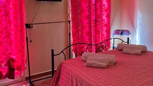 A hotel room with a bed, pink curtains, a TV mounted on the wall, and a chair. - Korisia accommodation in villa delle canfore surrounded by an olive grove with sea view (AVOLA)