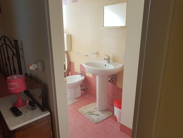 Un baño con lavamanos, bidé y inodoro. Hay un espejo sobre el lavamanos y un perchero en la pared.