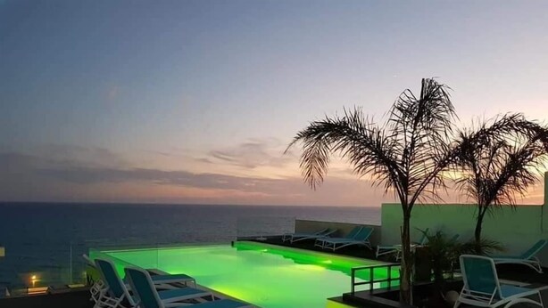 A rooftop infinity pool with green lighting, lounge chairs, and a view of the ocean at sunset.