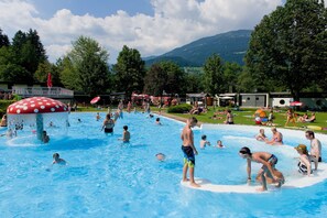 Una piscina con gente disfrutando del agua, rodeada de árboles y montañas al fondo.