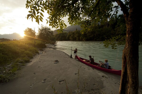 Orilla de río con una canoa y dos personas, una de pie y otra sentada, cerca de la orilla.