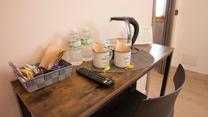 A wooden desk with a remote control, two mugs, two water bottles, and a basket of magazines. - Shardan Guest House (Alghero)