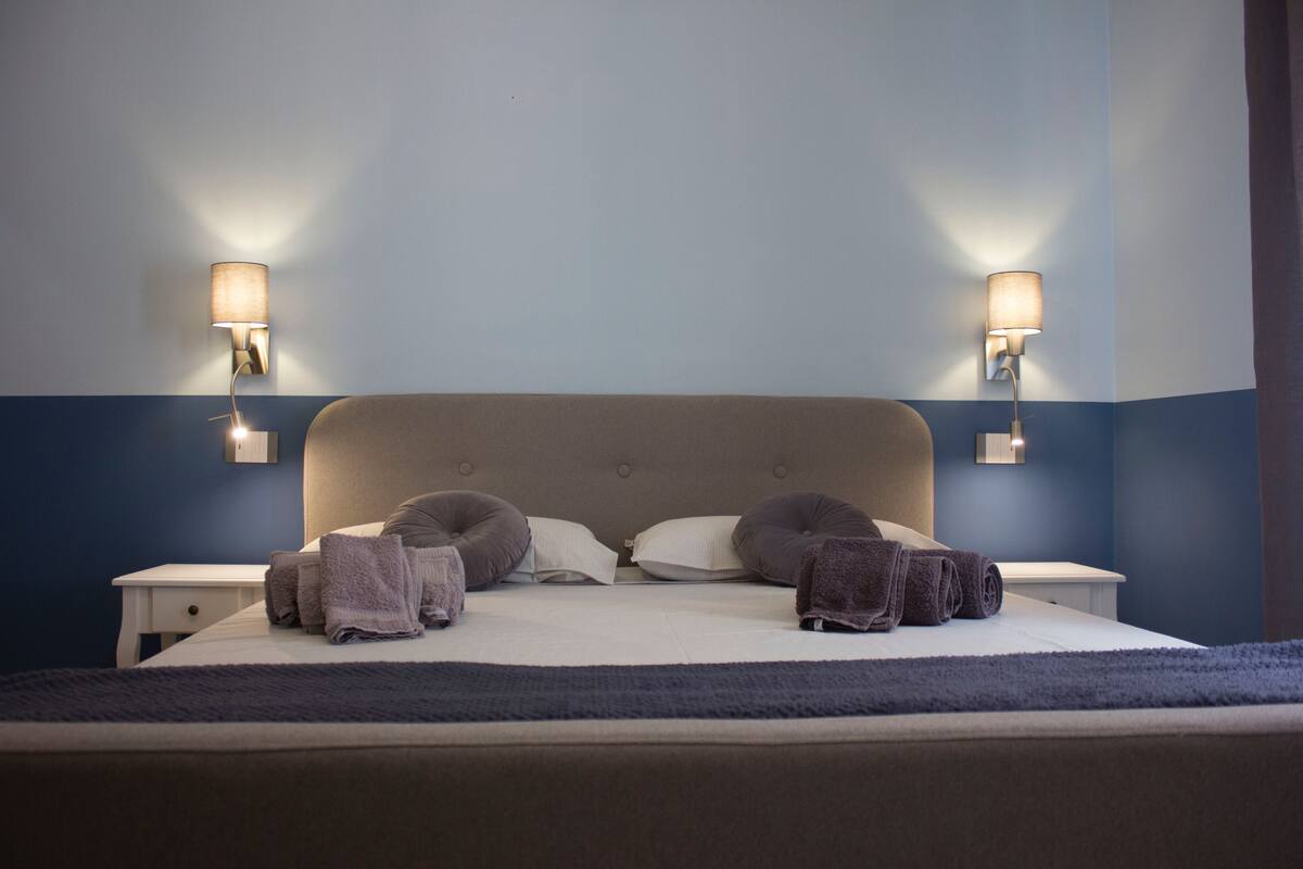 Una cama bien hecha con almohadas y toallas dobladas, dos mesitas de noche y lámparas de pared.