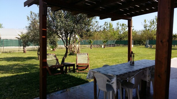 Ein Außenbereich mit einer überdachten Sitzgelegenheit, einem Tisch mit Tischdecke und Blick auf einen eingezäunten Garten mit Bäumen und einem Gebäude im Hintergrund.