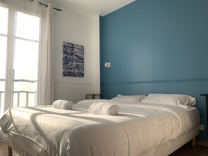 Um quarto com uma cama, roupa de cama branca, uma parede de destaque azul e um pôster com nuvem de palavras.