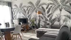 A living room with a grey sofa, a dining table with chairs, a TV on a stand, and a wall with a tropical leaf pattern. - Disneyland Paris 3 minutes away, sleeps 8, 2 bedrooms. (Chessy)