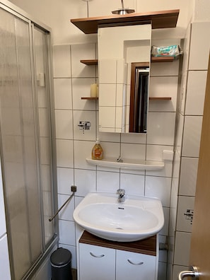 Un baño con un lavabo blanco, un espejo encima y una puerta de ducha. Hay estantes de almacenamiento sobre el espejo y un cesto de basura junto al lavabo.