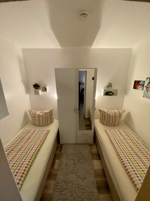 Habitación pequeña y luminosa con dos camas individuales, un armario, un espejo y una estantería con plantas.