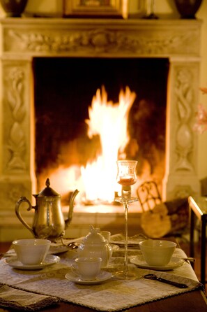Une table dressée avec des tasses à thé, une théière et un verre, placée devant une cheminée où l’on voit des flammes.