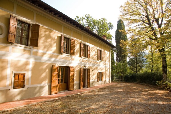 Un bâtiment à deux étages, doté de nombreuses portes et fenêtres en bois, entouré d’arbres et bordé d’un chemin de gravier.