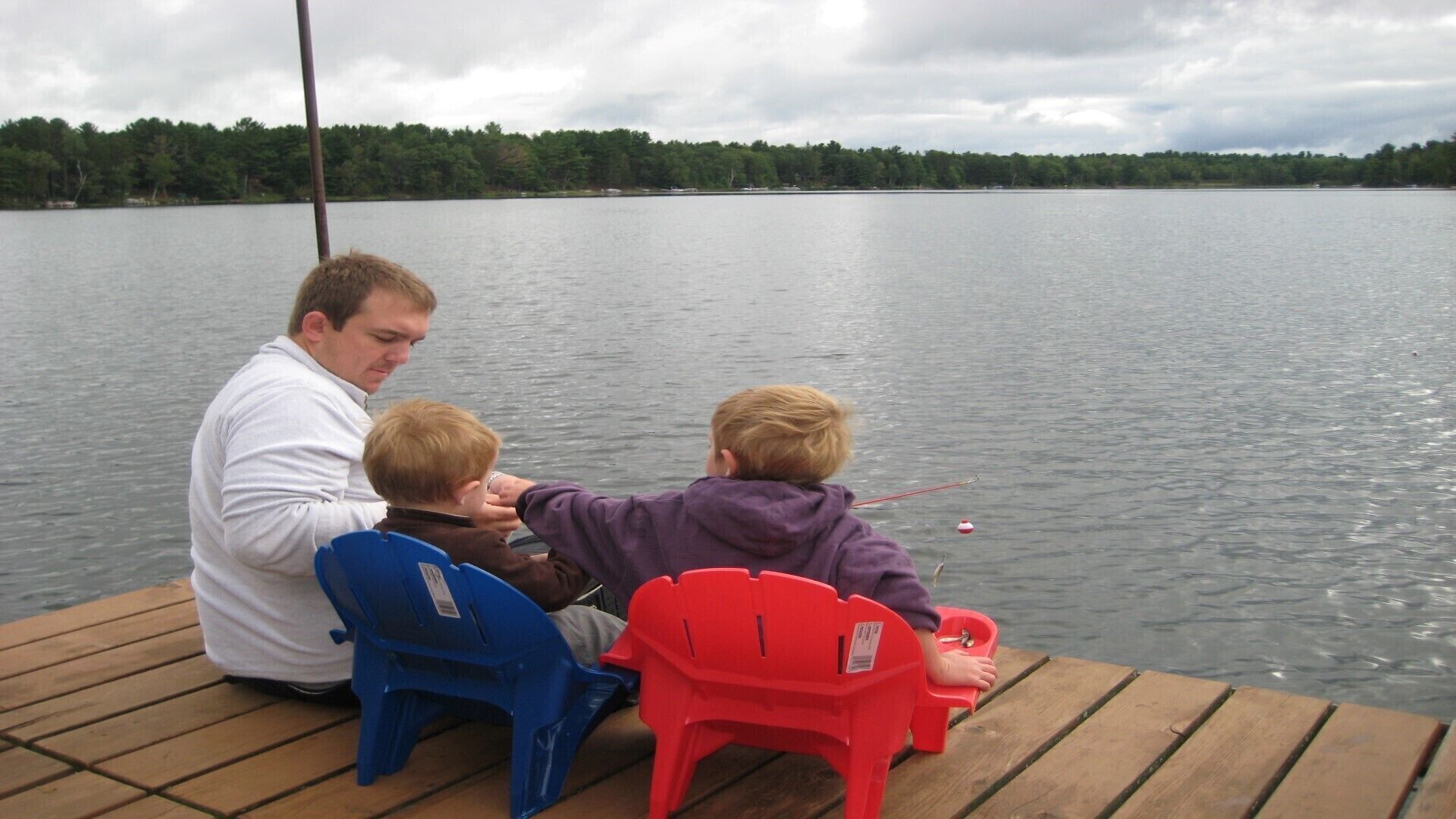A man and two children fishing on a wooden dock by a lake.
