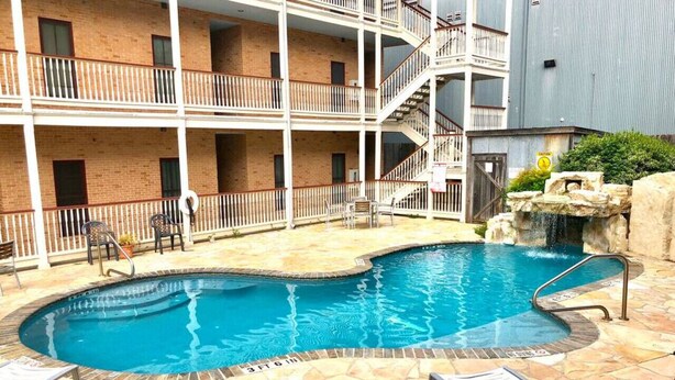 A swimming pool with a stone border, surrounded by lounge chairs and a multi-story building with balconies. - Long Weekend | Guadalupe Riverfront | Downtown Gruene | Pool (New Braunfels)