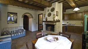 Un intérieur rustique avec une cheminée en pierre, des poutres en bois, une table à manger où se trouve un chat, et un canapé bleu.