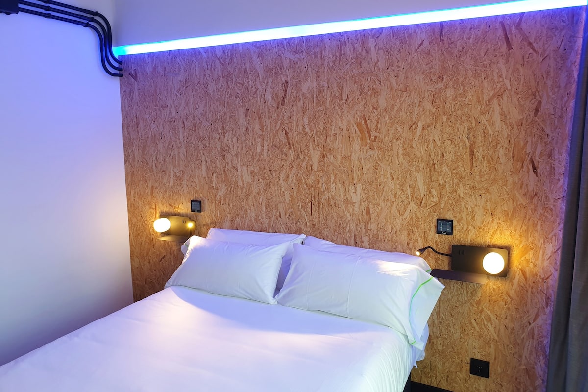 A neatly made bed with white linens in a room with wooden paneling on the walls and two wall-mounted light fixtures.