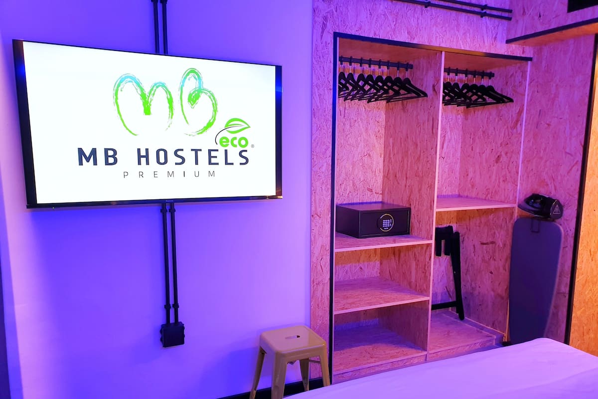 A room with a bed, a TV displaying 'MB HOSTELS PREMIUM' logo, a wardrobe with hangers, and a small stool.