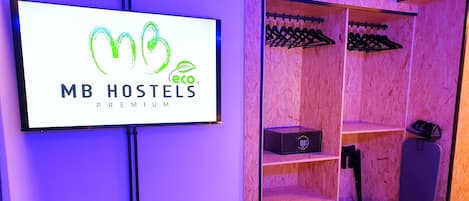 Una habitación con cama, televisor mostrando el logotipo de 'MB HOSTELS PREMIUM', armario con perchas y un taburete pequeño.