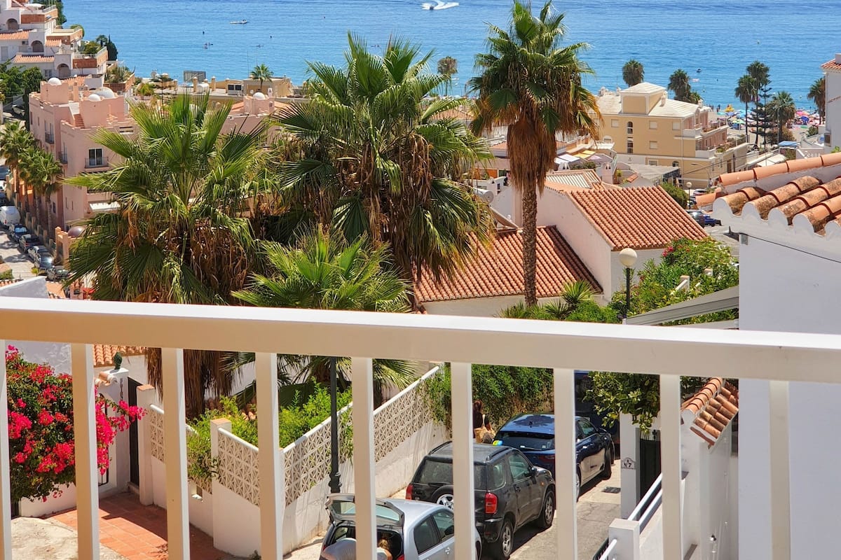 A coastal residential area with a view of the sea, palm trees, and a balcony overlooking the street.