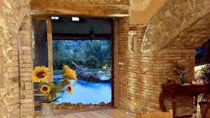 Zona pranzo con vista su piscina e giardino, pareti in pietra e tavolo in legno apparecchiato per due.