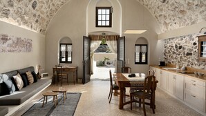 Un vaste espace de vie doté d’un plafond en pierre, d’une table à manger avec des chaises, d’un canapé avec des coussins et d’une vue sur un espace extérieur, accessible par une porte ouverte.
