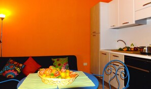 A compact kitchen with white cabinets, a sink, and a countertop with a pot and vegetables. A dining table with a basket of fruit is in the foreground.