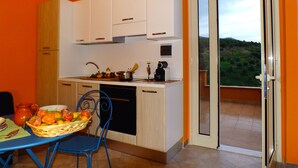 Cocina compacta con armarios blancos, una mesa de comedor con sillas y vistas a un paisaje verde a través de una puerta corrediza de vidrio.