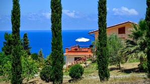 Una casa costera con terraza, rodeada de árboles altos y vista al mar.