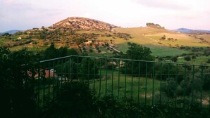 A hilly landscape with a prominent hill in the background, a fenced area in the foreground, and scattered buildings. - Nunziata House (Mineo)