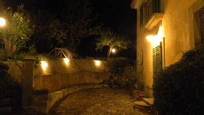 A stone pathway leading to a building with a curved wall and a small garden area with potted plants and a bench. - Nunziata House (Mineo)