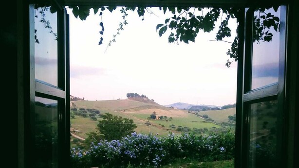 A stone pathway leading to a glass door opening to a scenic view of a hilly landscape with a distant hill and flowering plants. - Nunziata House (Mineo)