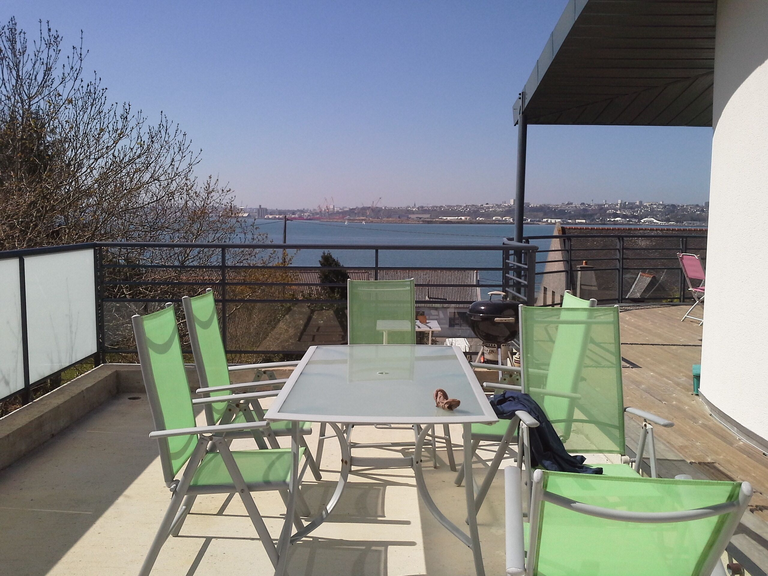 Een terras buiten met een tafel en stoelen, met uitzicht op een waterpartij en een verafgelegen stadsgezicht.