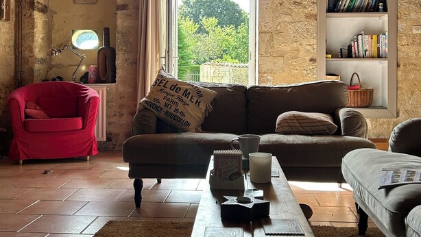 Una habitación rústica con vigas de madera a la vista, un sillón rojo, un sofá, una estantería y un ventanal con vistas al verdor.