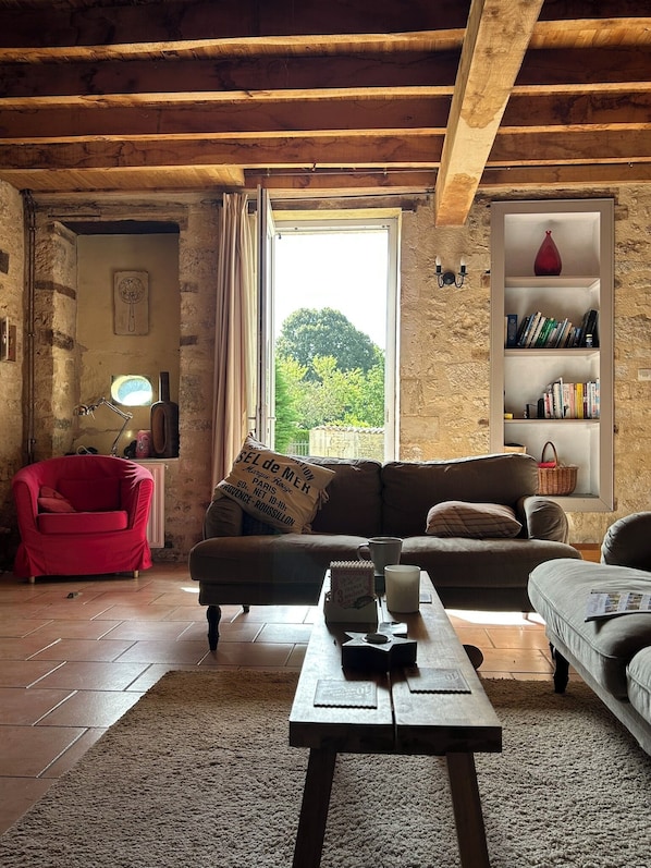 A rustic room with exposed wooden beams, a red armchair, a sofa, a bookshelf, and a window with a view of greenery.