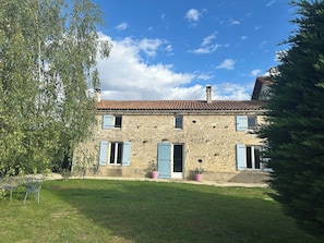 A stone house with a tiled roof, two blue doors, and a small garden with a tree and a table.