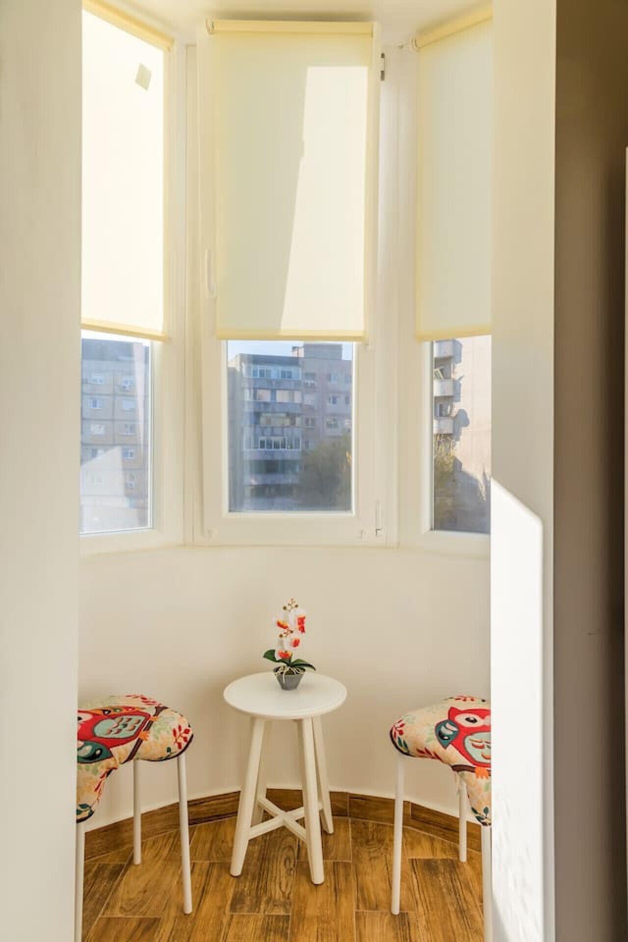 Ein Eckzimmer mit Erkerfenster, ein runder Tisch mit einem Blumentopf und zwei Stühle mit bunten Mustern.