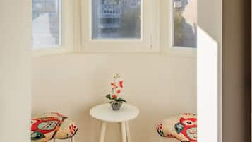 Ein Eckzimmer mit Erkerfenster, ein runder Tisch mit einem Blumentopf und zwei Stühle mit bunten Mustern.