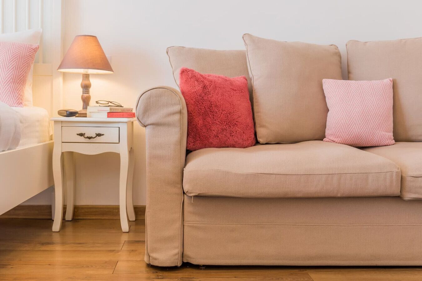Ein beiges Sofa mit zwei Kissen, ein weißer Nachttisch mit einer Lampe und ein Kissen mit Muster.