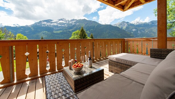 Una terraza de madera con sofá y muebles de mimbre, con vistas a un paisaje montañoso.