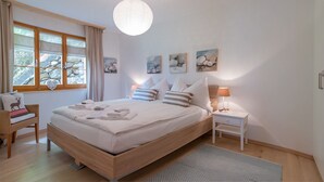 Una cama bien hecha con almohadas y toallas dobladas, una mesita de noche con una lámpara, una silla con cojín y una ventana con cortinas.