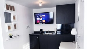 Een kleine keuken met een inbouwkoelkast, een koffiezetapparaat en een spoelbak. Een flatscreen televisie hangt boven het aanrecht en toont diverse streamingdiensten.