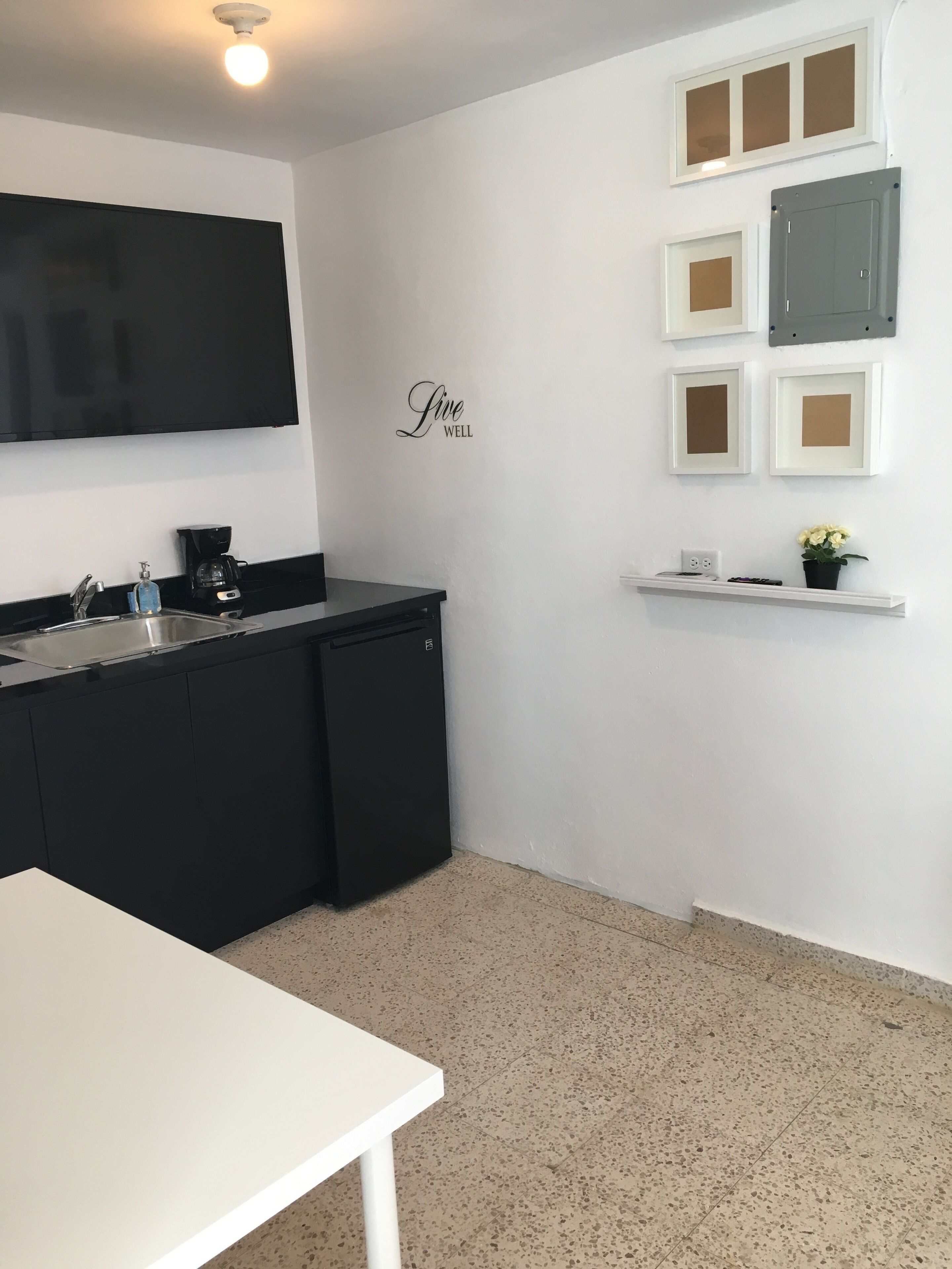 A modern kitchen with a black countertop, a white sink, and a black refrigerator. There is a white shelf with a potted plant and a framed picture.