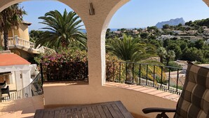 Ein Balkon mit Holztisch und Stuhl, Blick auf eine Küstenstadt und ein Berg in der Ferne.