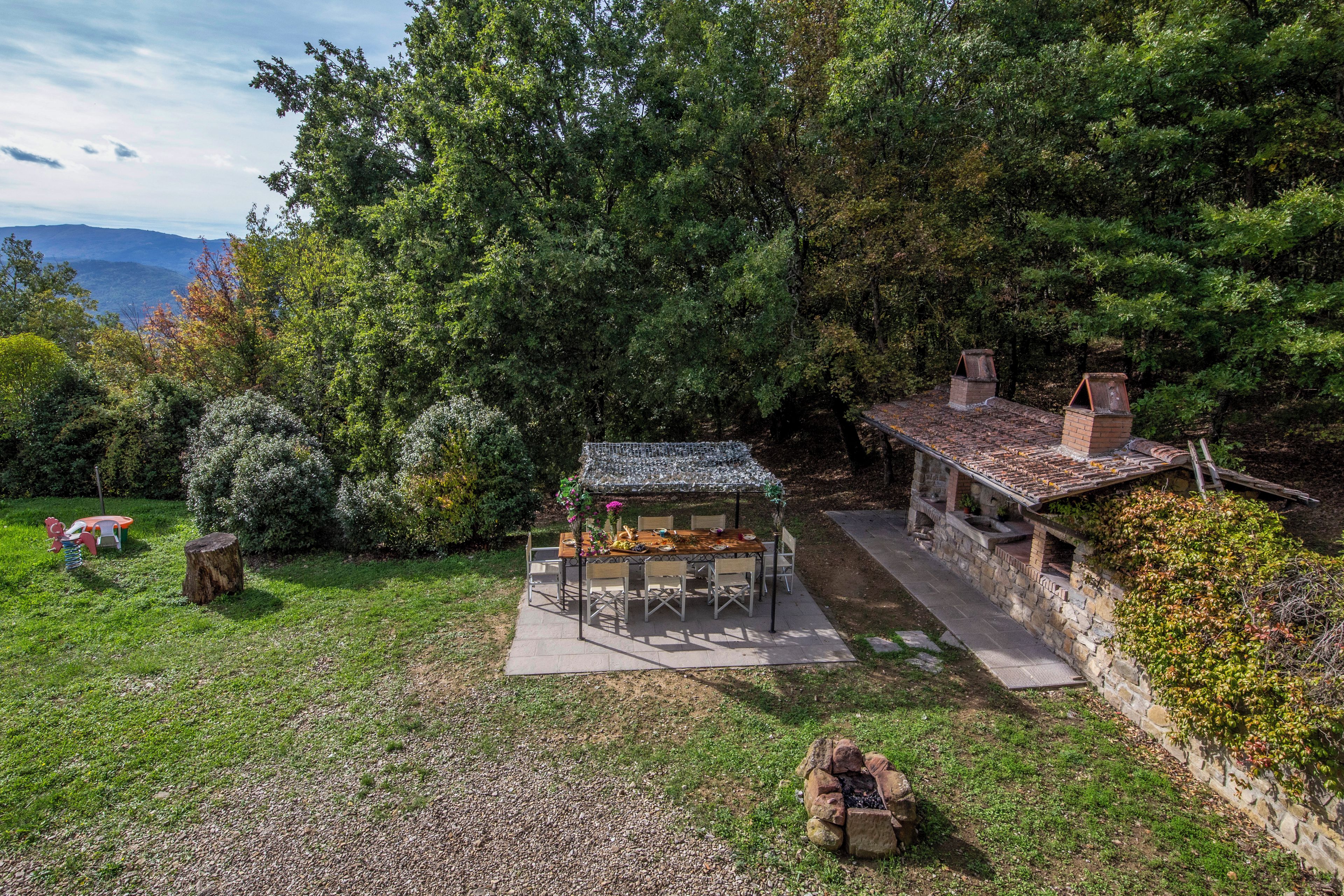 Un espace de restauration extérieur rustique, avec une table et des chaises, entouré d’arbres et d’un mur de pierres.