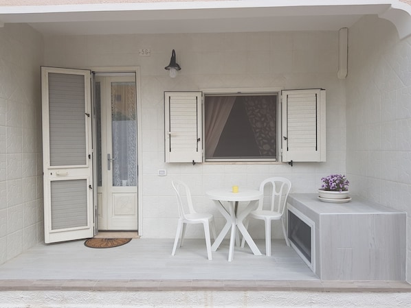 A small patio with a white door, two white chairs, a small table, and a potted plant.