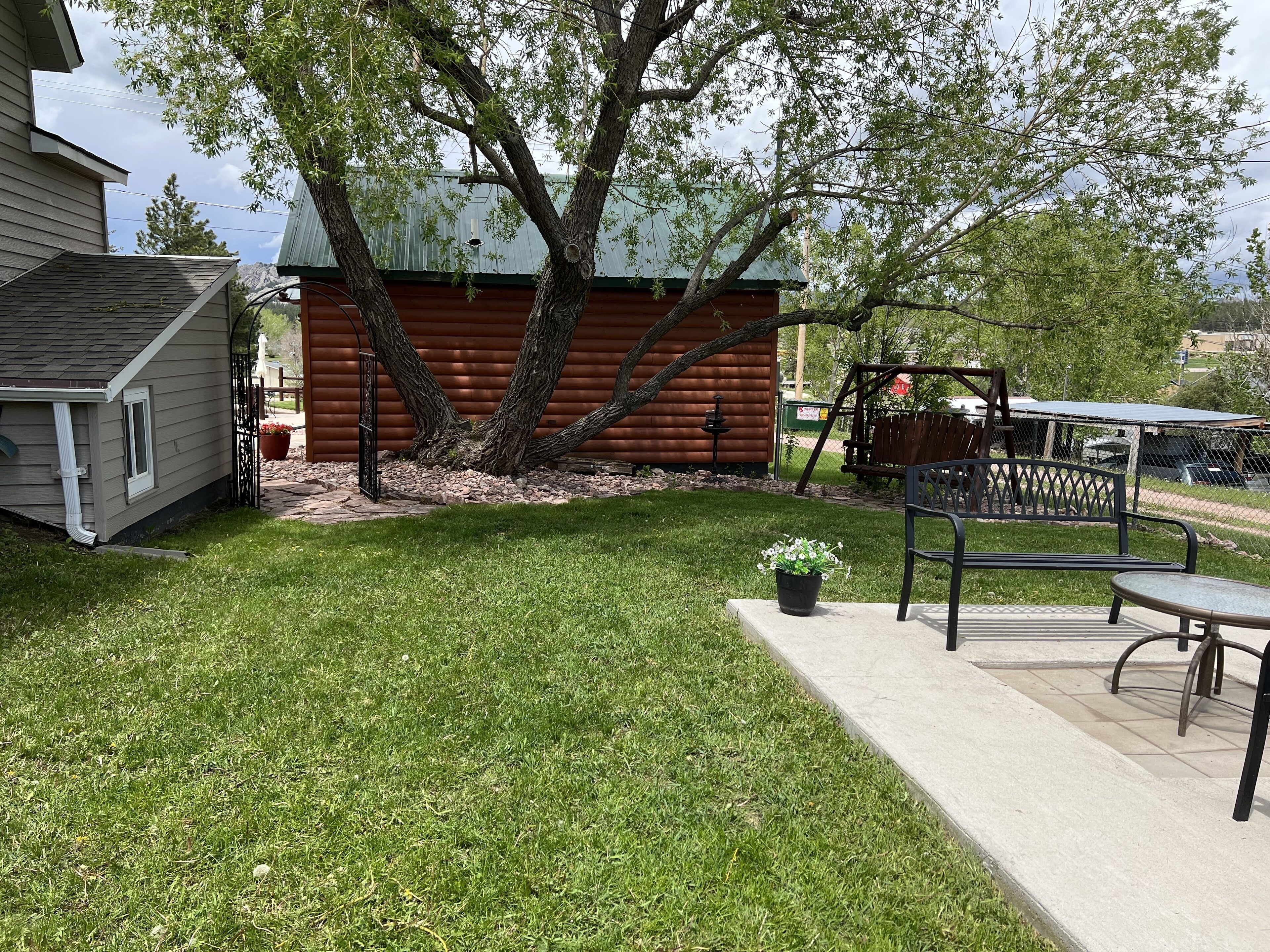 A backyard with a wooden cabin, a swing set, and a patio area with a table and chairs.